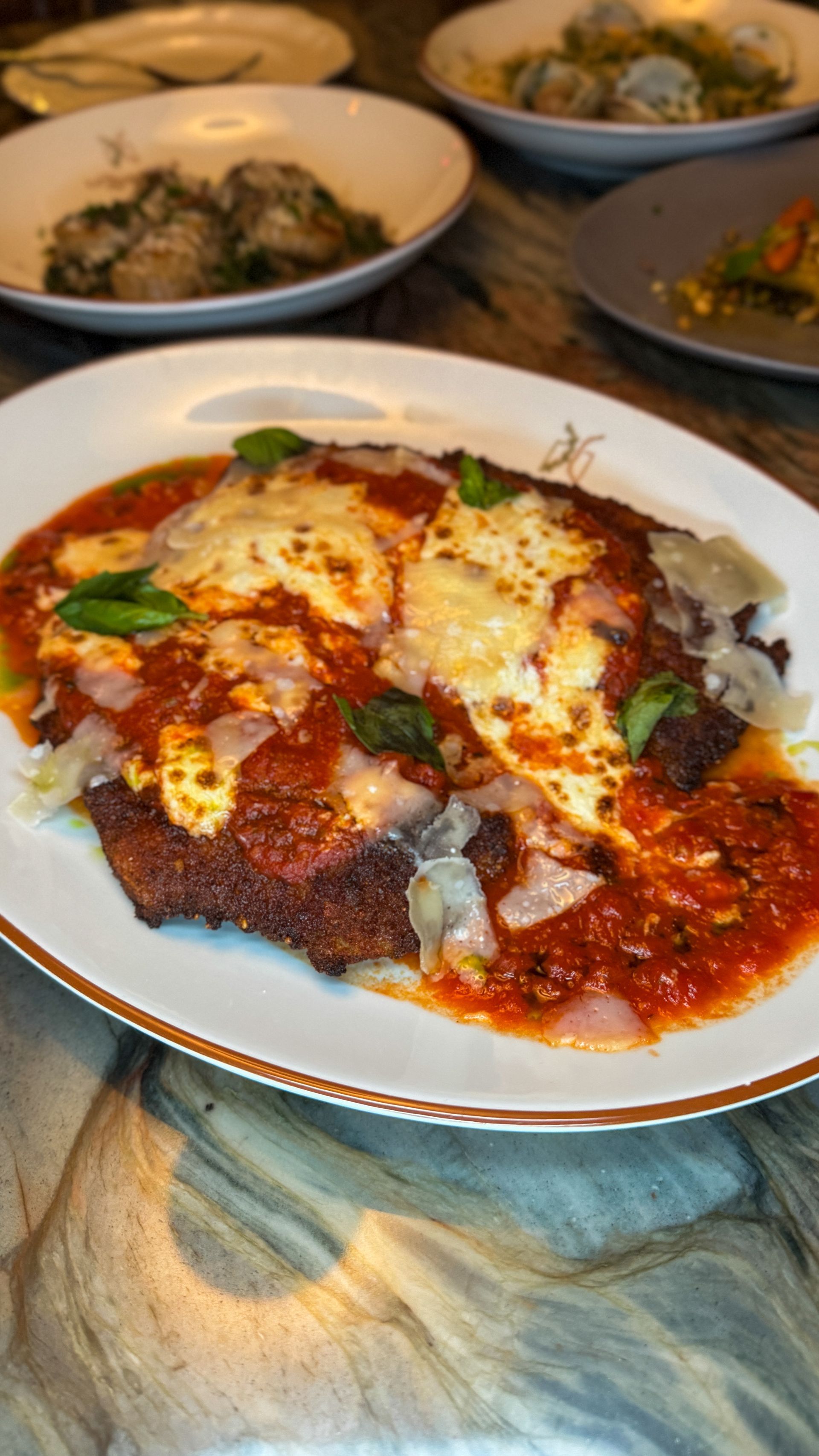Chicken parmesan on a white plate with tomato sauce and melted cheese, basil garnish. Other plates in the background.