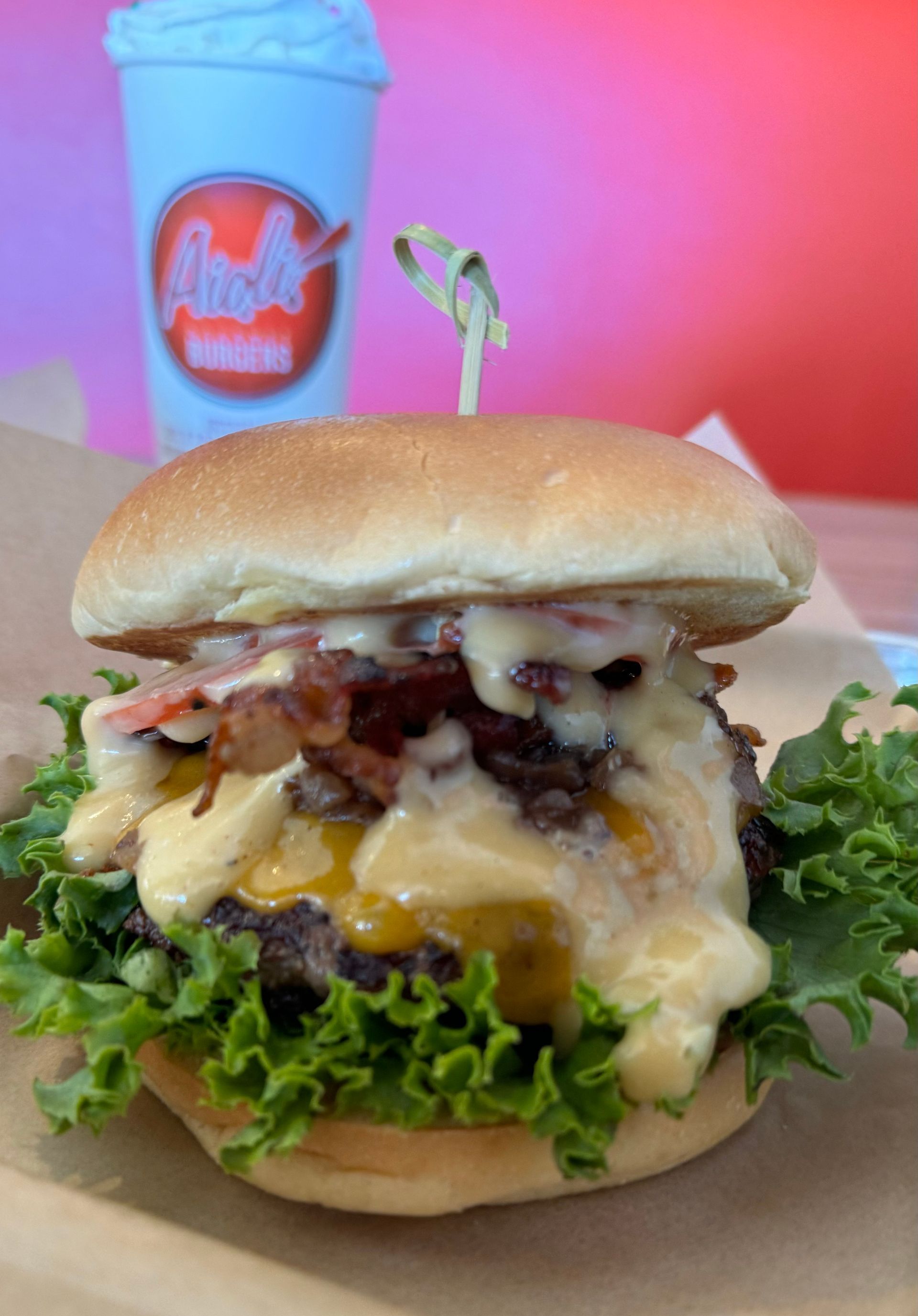 Aioli Gourmet Burgers - A hamburger with lettuce , cheese , bacon and a drink in the background.