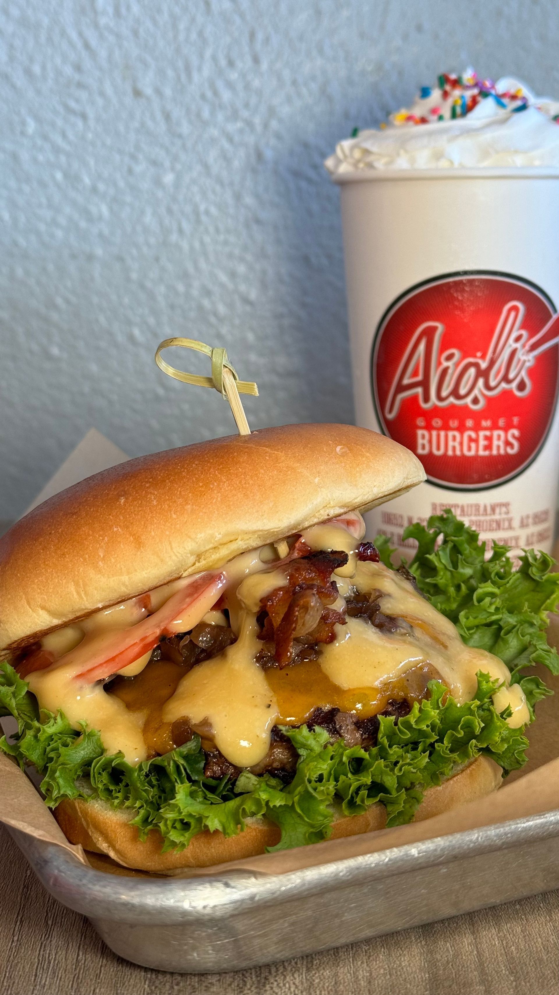 Aioli Gourmet Burgers - A hamburger is sitting on a metal tray next to a cup of milkshake.