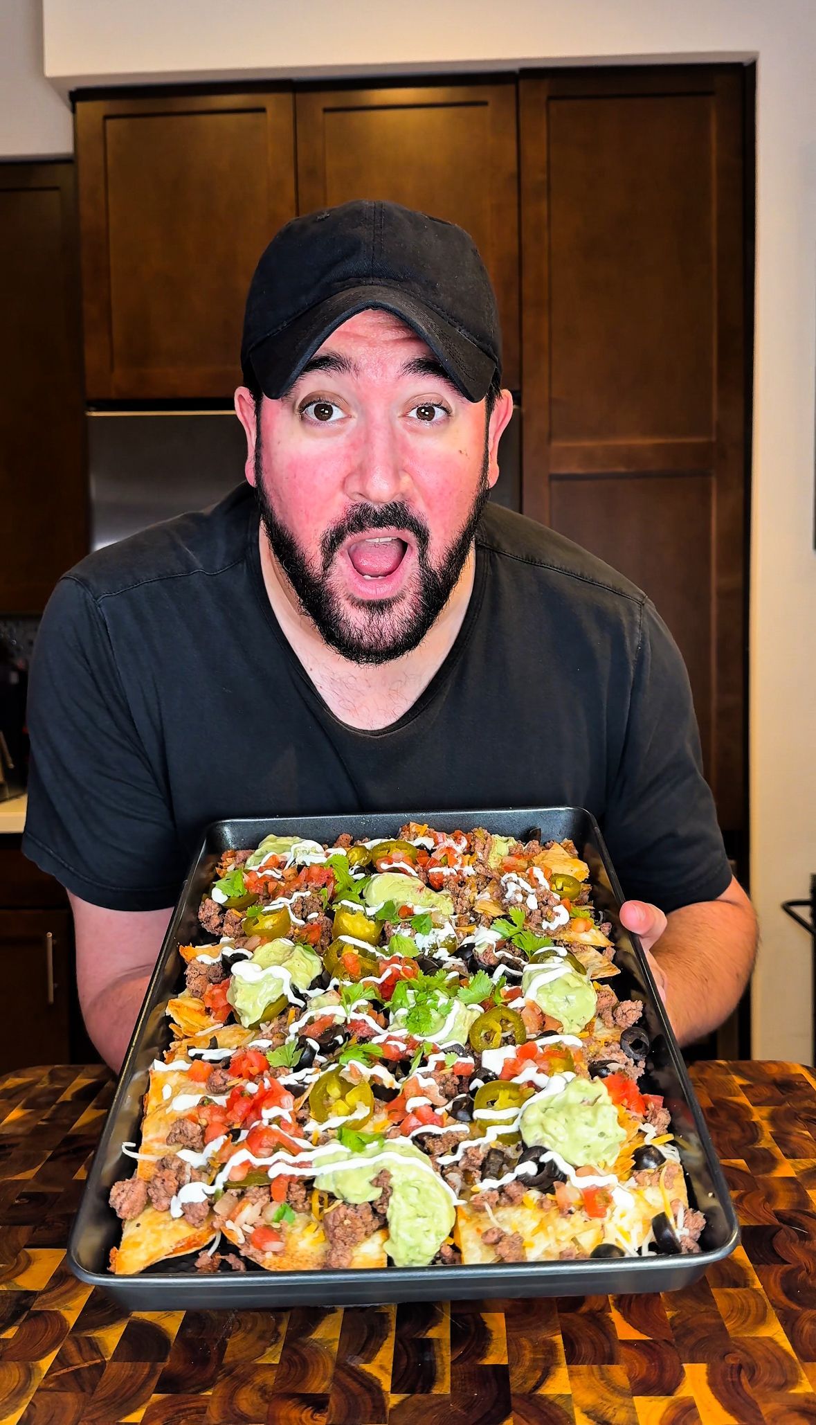 Man holding a tray of loaded nachos, brown cabinets in background, excited expression.