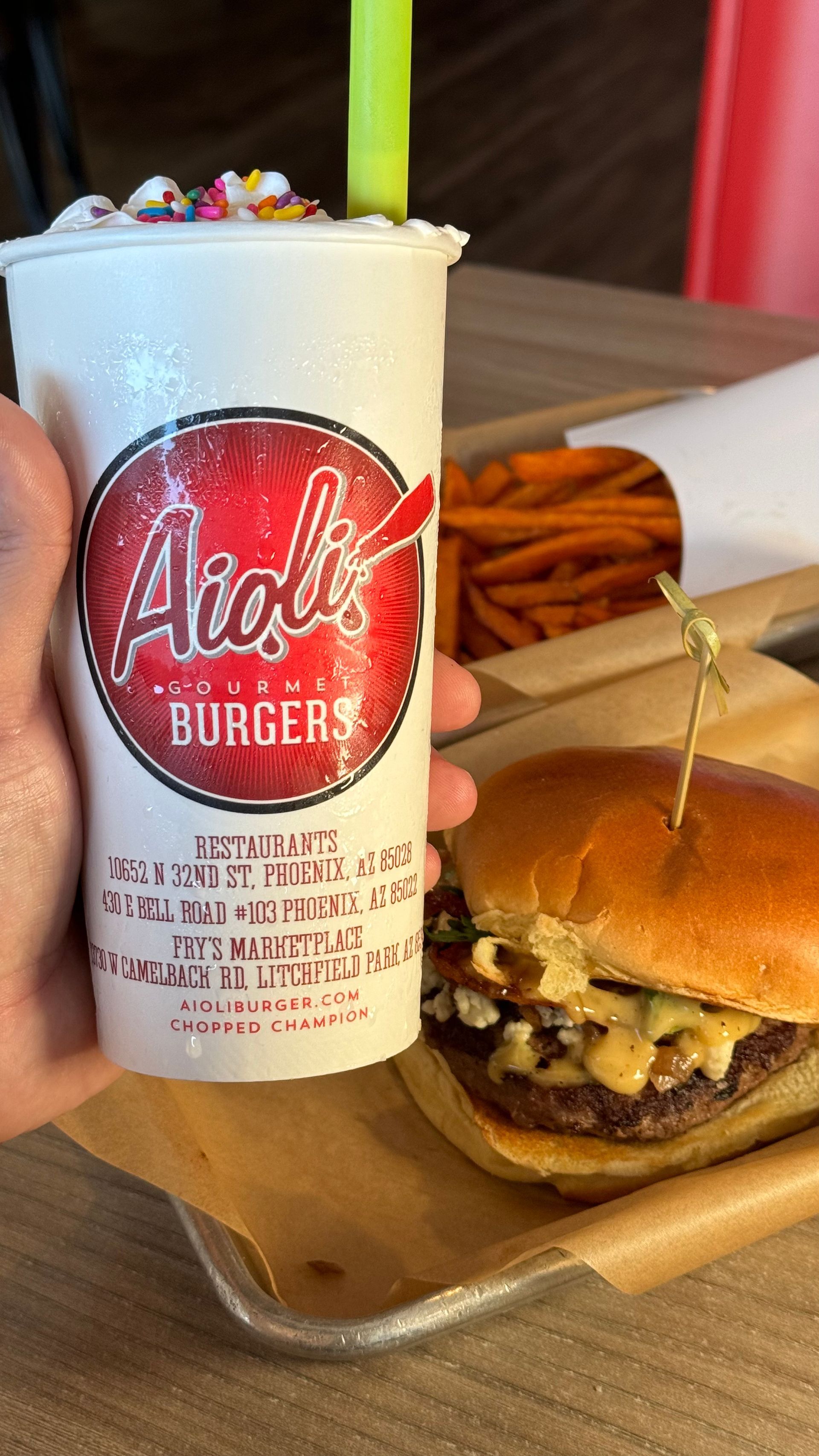 Aioli Gourmet Burgers - A person is holding a cup of milkshake next to a hamburger.