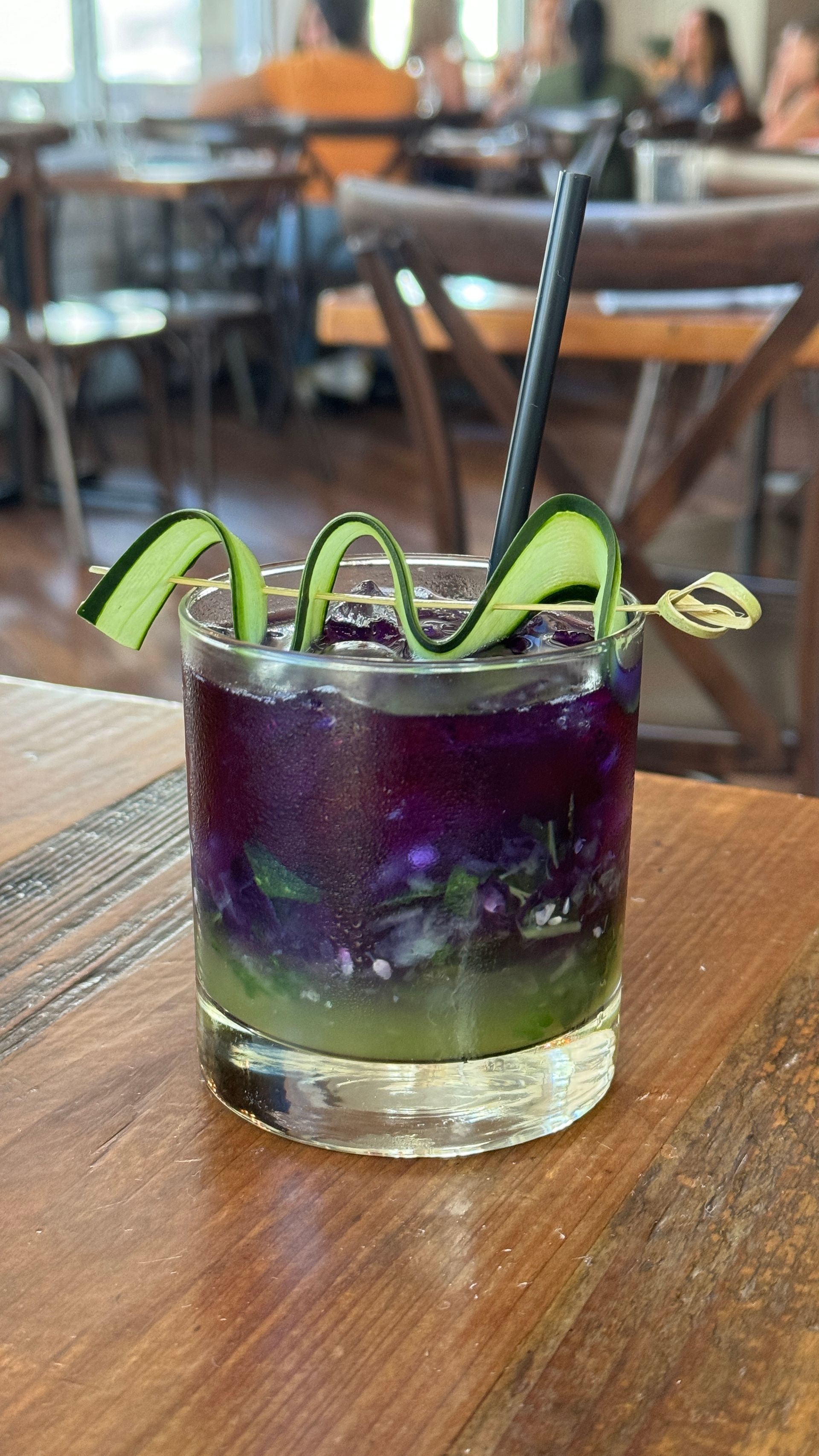 A purple and green cocktail with a cucumber garnish is on a wooden table.