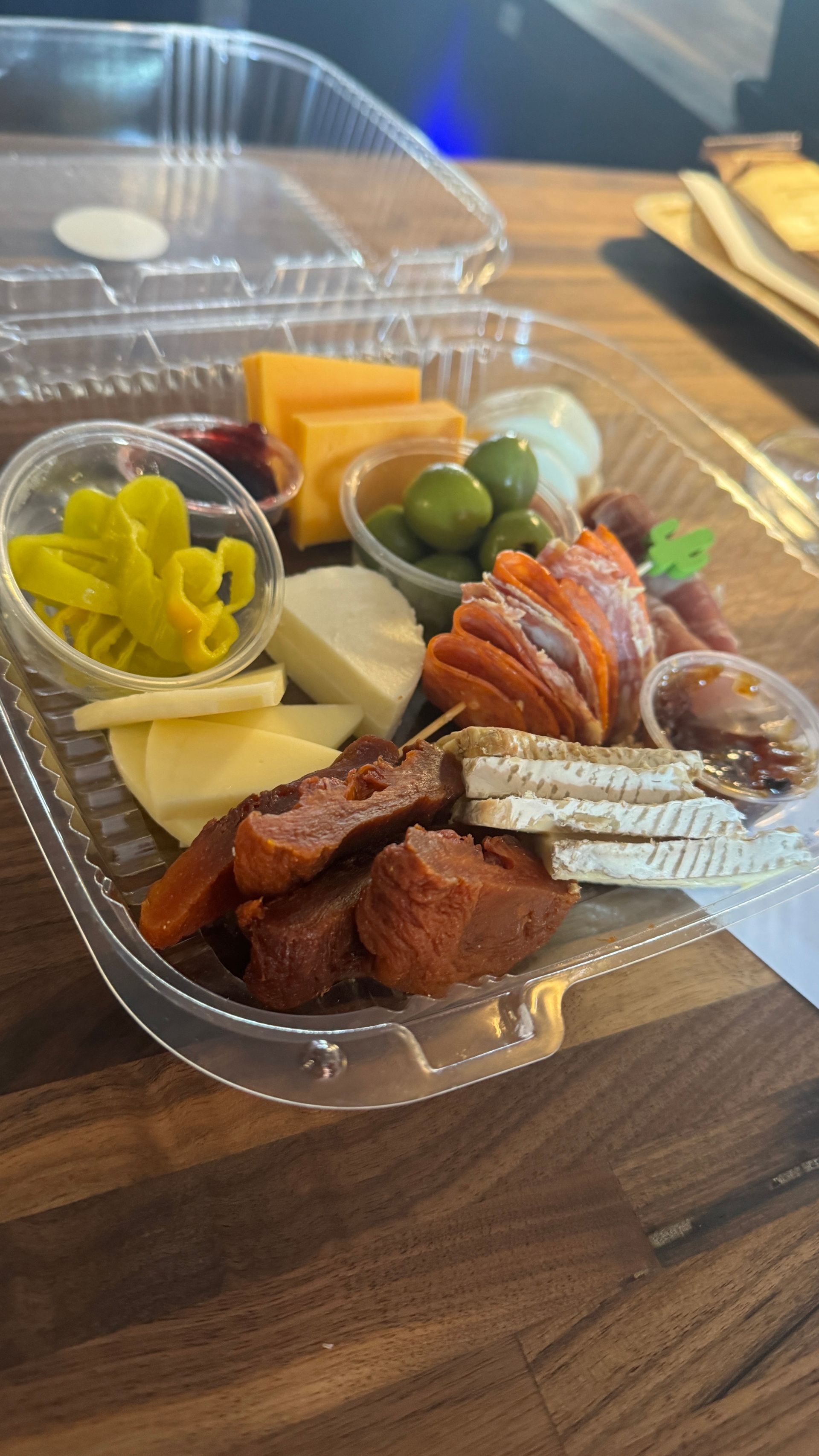 A plastic container filled with meat , cheese , olives and pickles on a wooden table.