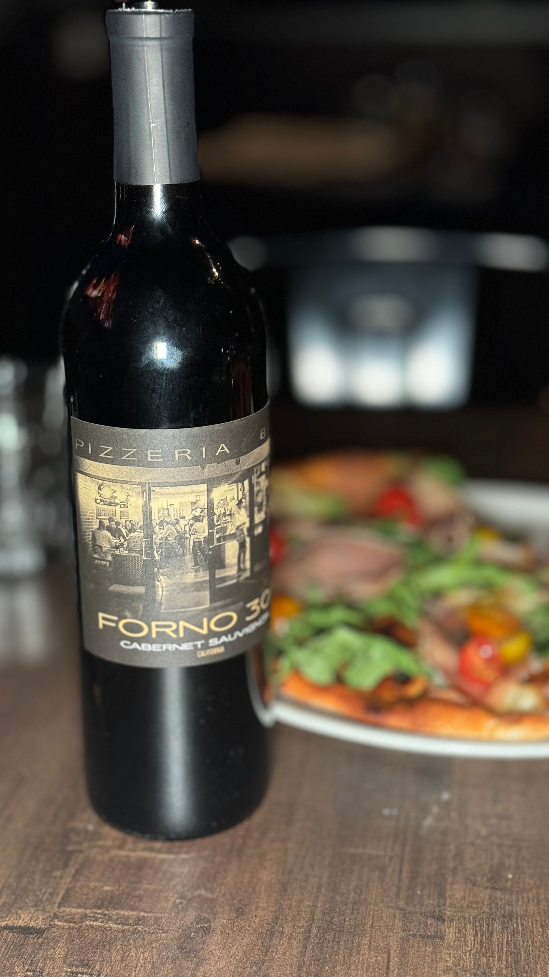 A bottle of wine is sitting on a wooden table next to a pizza.