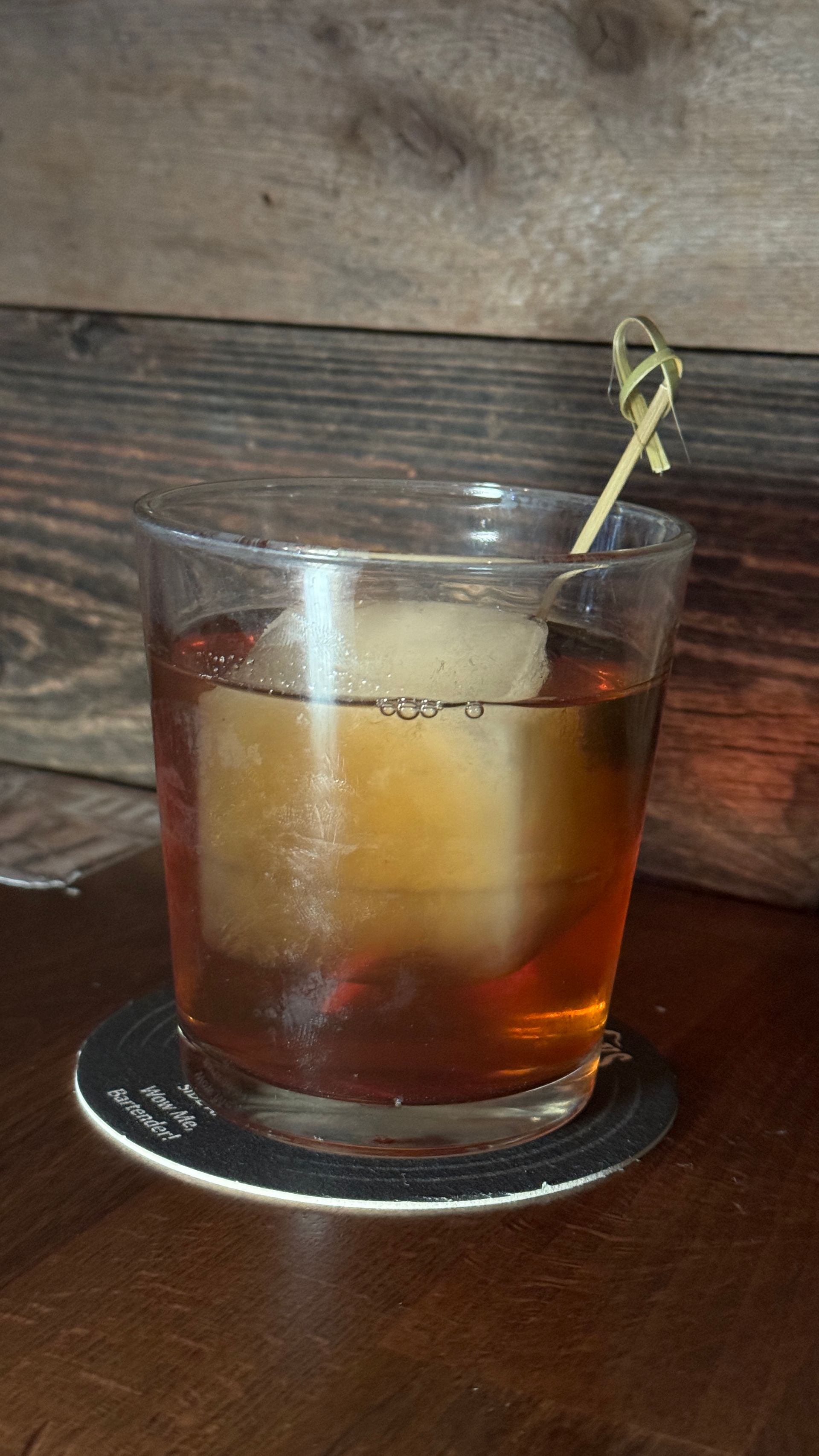 A glass of whiskey with a toothpick in it is on a coaster on a wooden table.