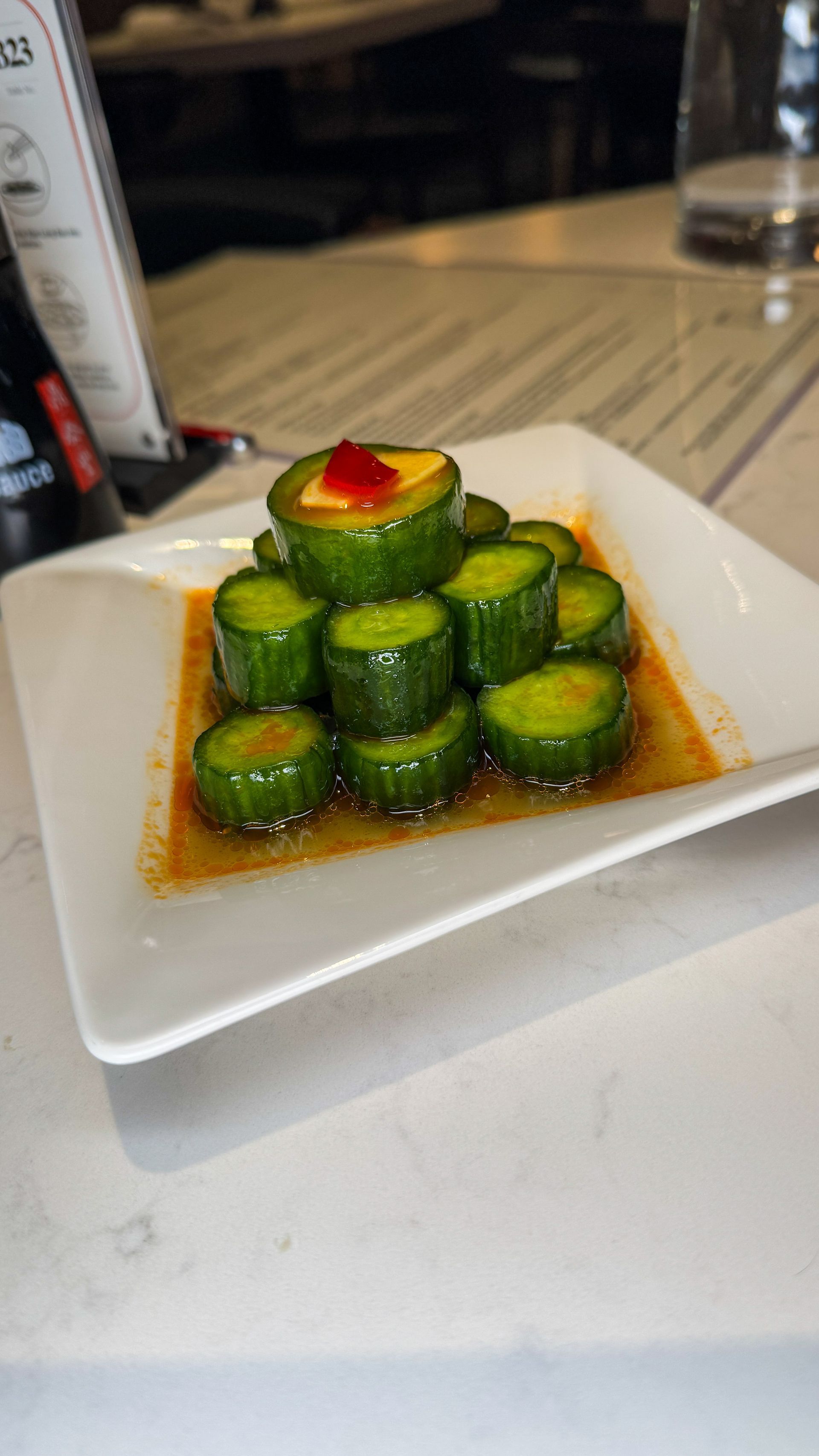 A small white square plate holds a stack of sliced cucumbers in a spicy, reddish-brown dressing with a red chili topper.