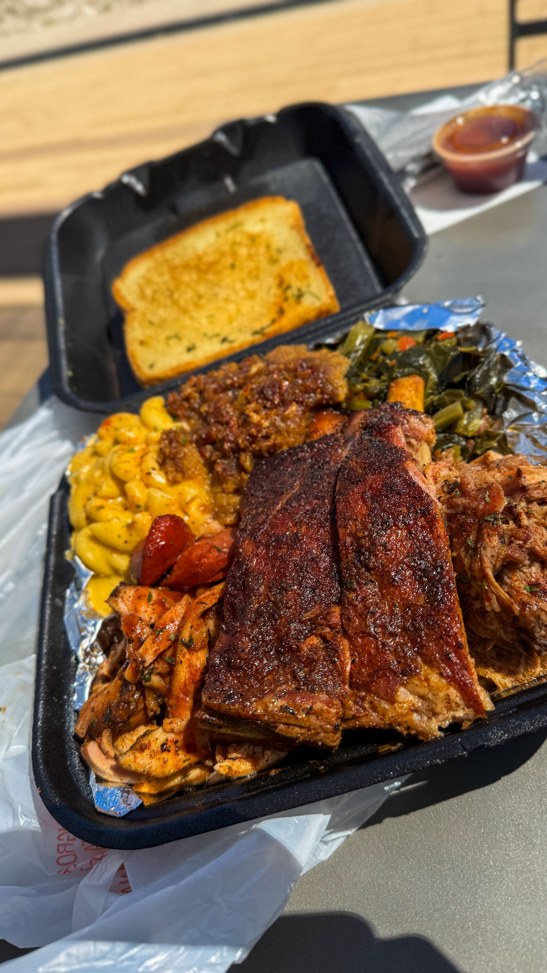 A takeout tray filled with barbecue ribs, mac and cheese, collard greens, and pulled pork, with a side of garlic bread.