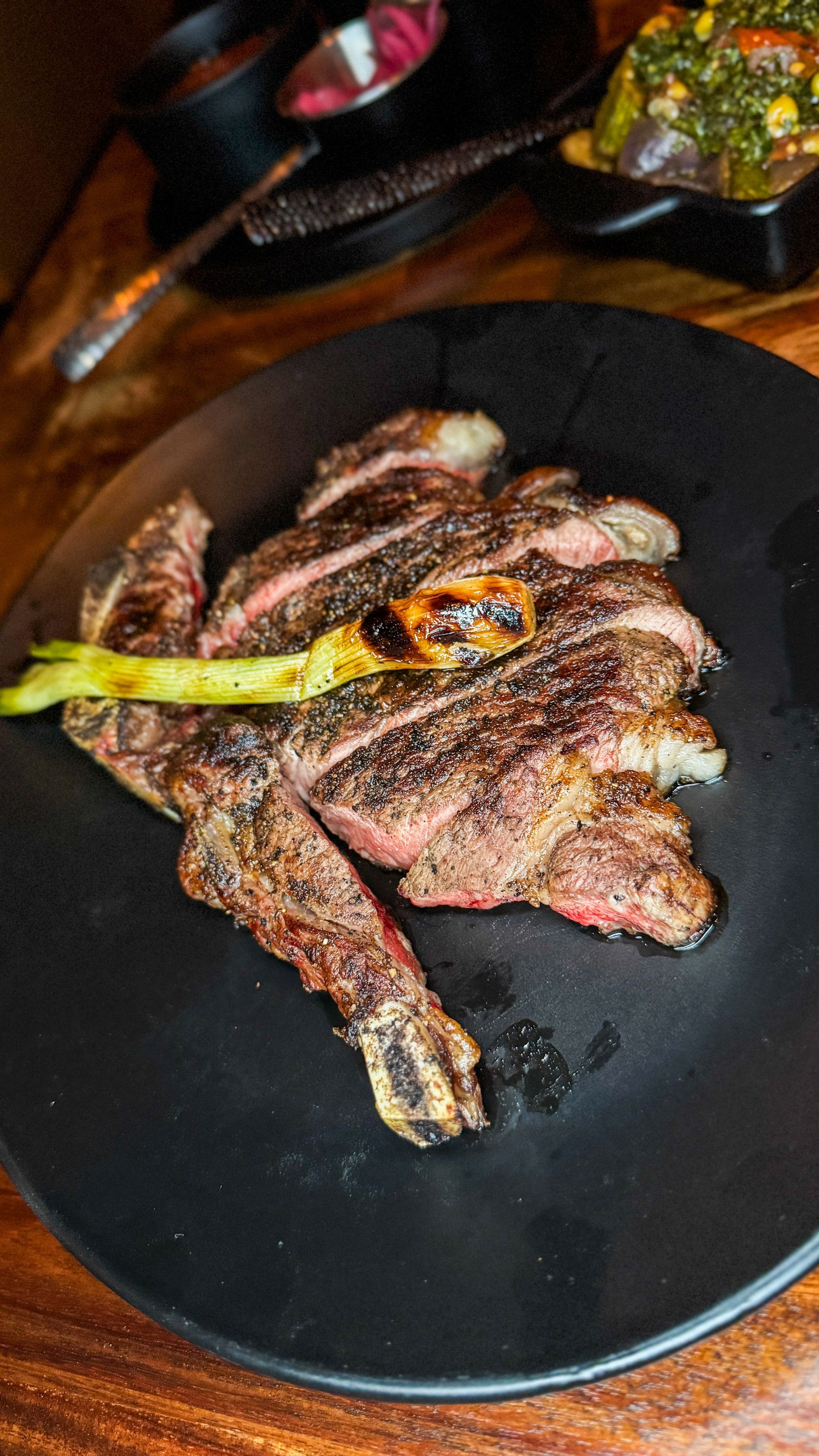 Sliced grilled steak served on a black plate with a charred scallion, accompanied by side dishes on a wooden table.