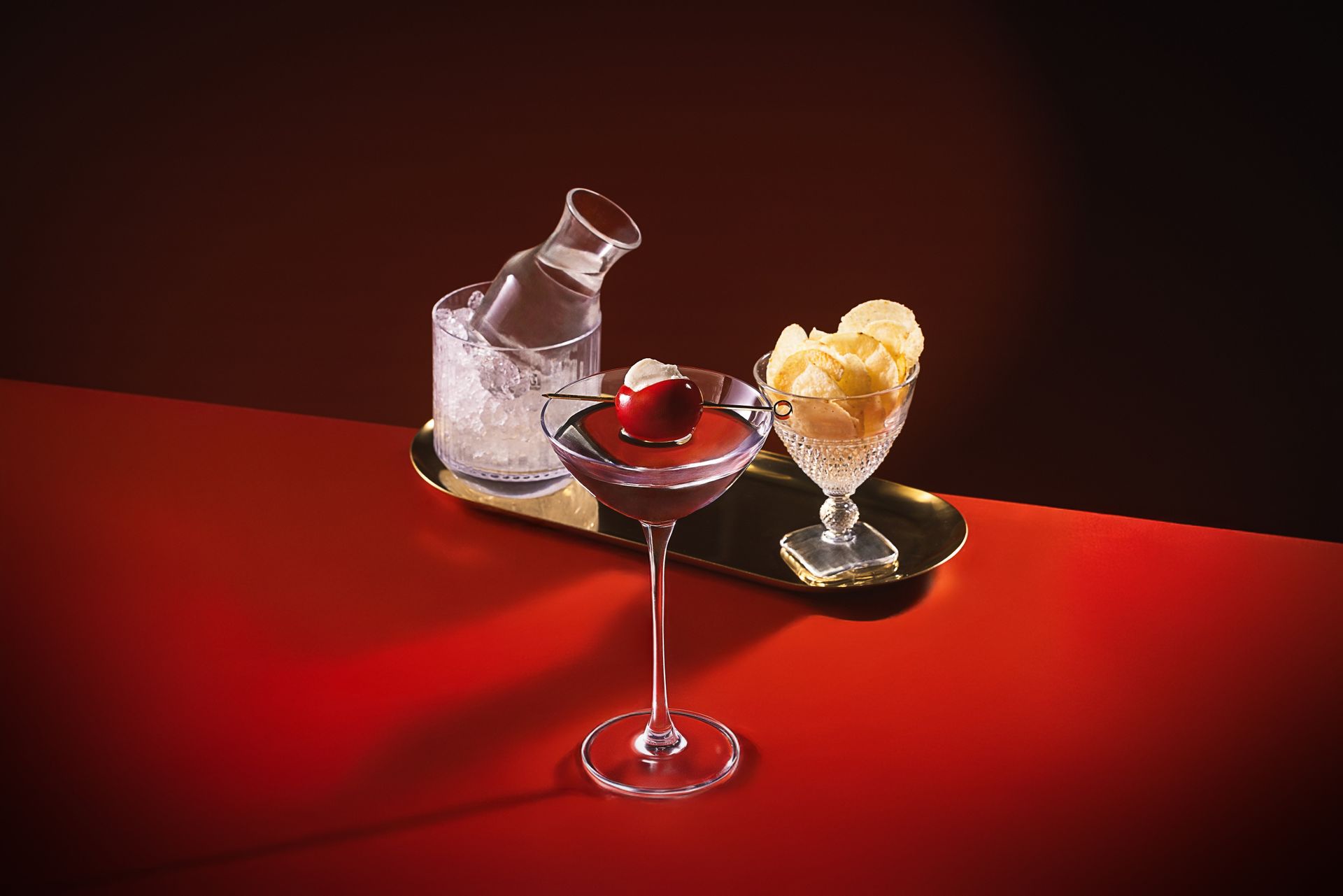 A martini glass with a cherry in it is on a table next to a bowl of chips.