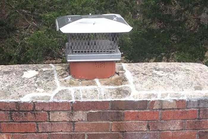 Chimney with metal cap and screen on a brick structure against a backdrop of green foliage.