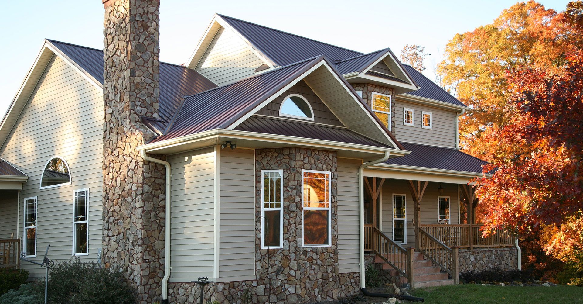 House with brown metal roof, stone chimney, tan siding, and autumn trees.
