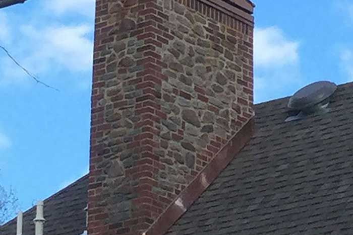 Brick and stone chimney on a dark shingled roof against a partly cloudy blue sky.