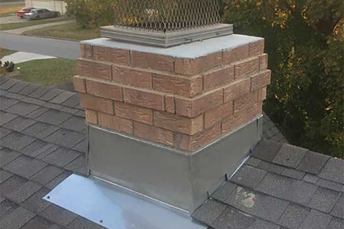 Brick chimney with metal flashing on a gray shingle roof, topped with a chimney cap.
