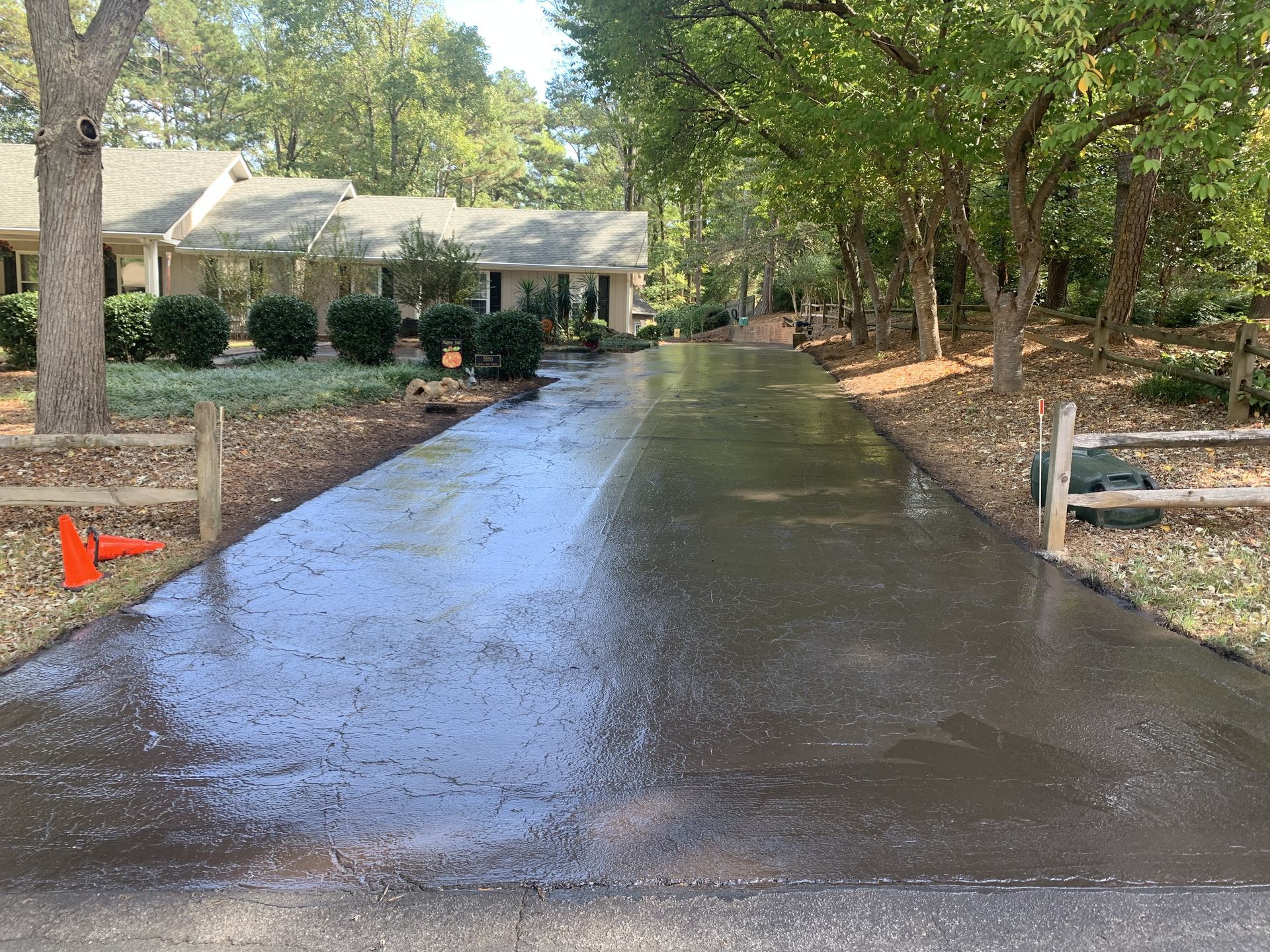 A long, wet asphalt driveway leading to a suburban house, flanked by green trees and wooden split-rail fences.