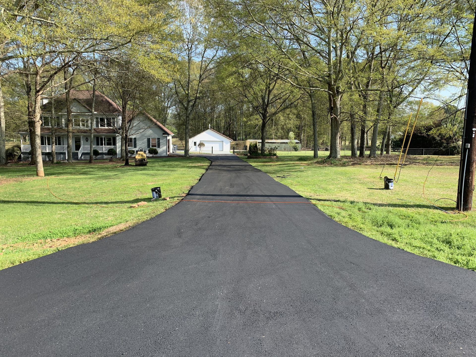 A freshly paved black asphalt driveway leads toward a two-story white house and a small shed on a grassy, tree-lined lot.