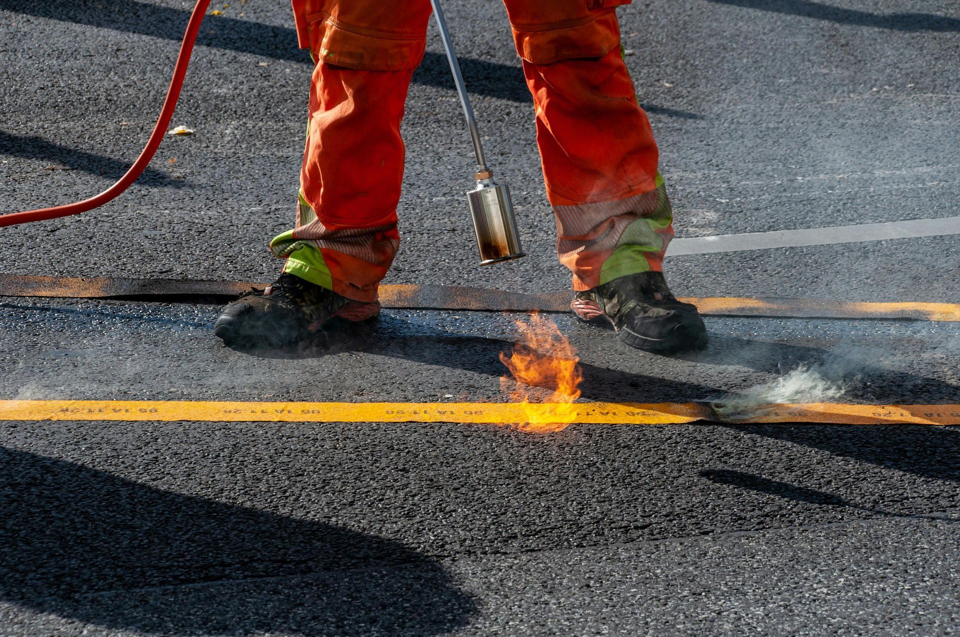 A worker in orange trousers uses a propane torch to melt yellow thermoplastic road marking onto black asphalt.