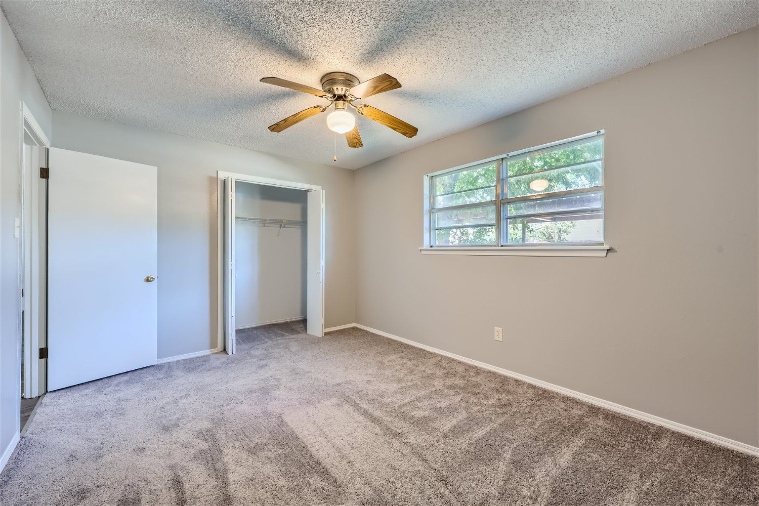 Empty bedroom with neutral walls, carpet, and a ceiling fan; closet and window visible.