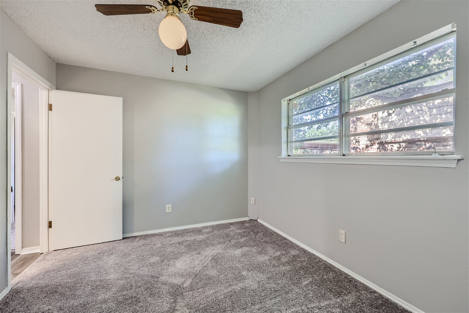 Empty gray bedroom with a closed white door, window, ceiling fan, and carpeted floor.