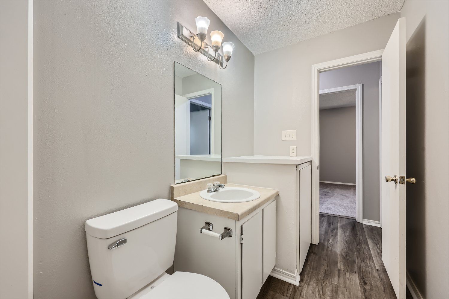 Bathroom with a white toilet, vanity, mirror, and open door to a hallway with wood-look flooring. Gray walls.