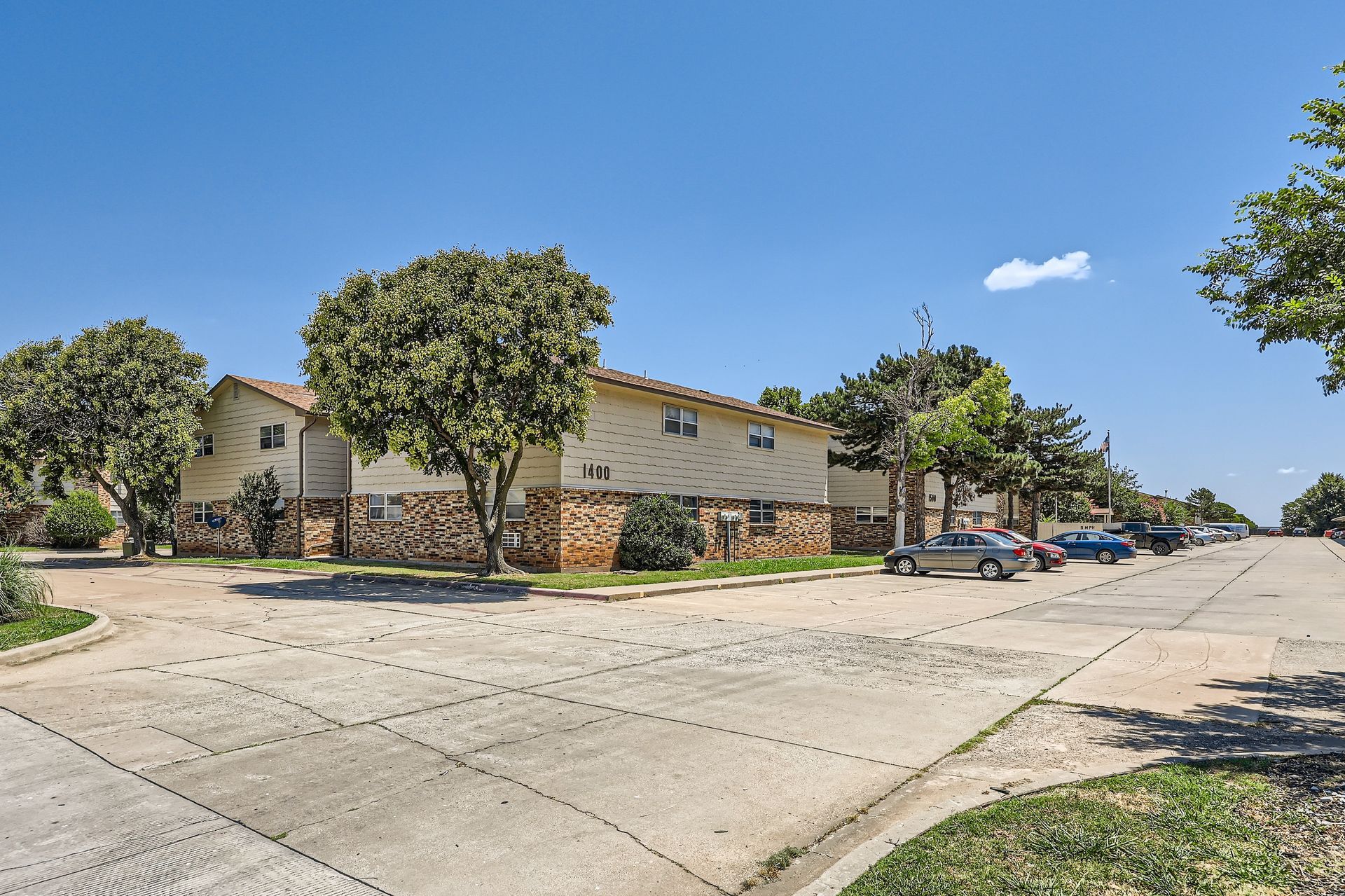 Apartment buildings with tan siding and stone facade on a sunny street with parked cars and trees.