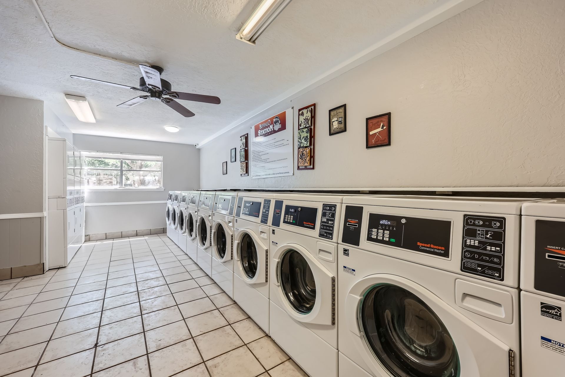 Laundry room with multiple washing machines in a row; tiled floor, ceiling lights, and framed posters on the wall.