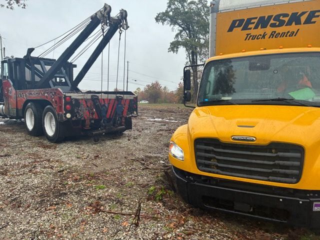 A bright yellow Penske rental truck parked on a muddy, leaf-covered field next to a large red tow truck.