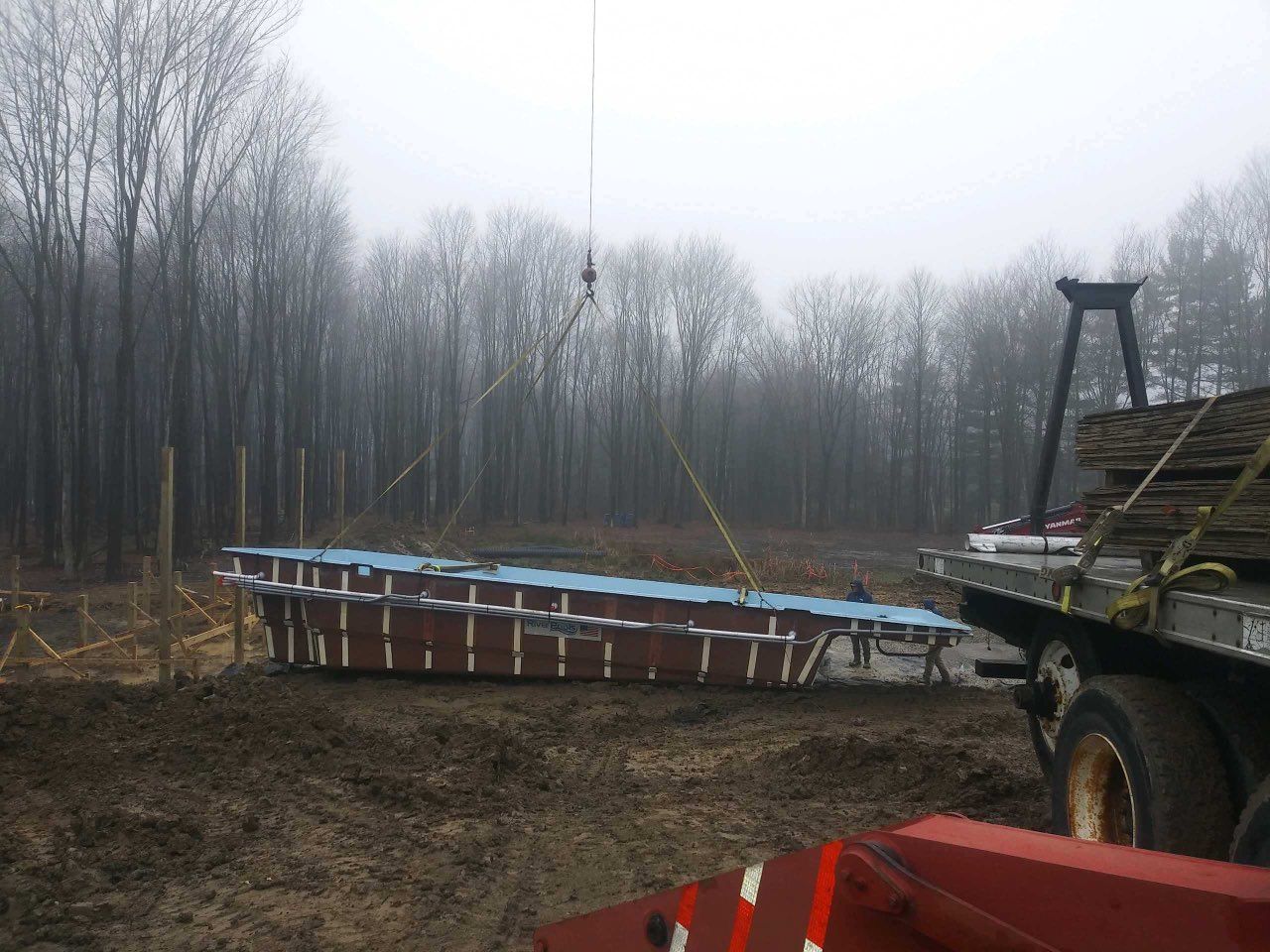 A crane lifting a large, rectangular brown pool shell into a muddy construction site, with a truck nearby .