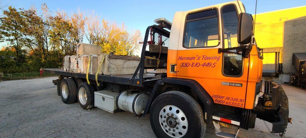 An orange and white flatbed truck parked on a gravel lot, loaded with concrete blocks secured by yellow straps.
