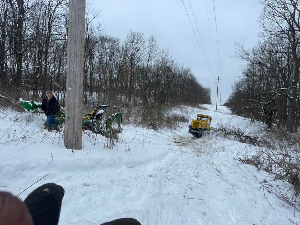 Snowy road with a small yellow vehicle, two people near fallen green equipment, and utility poles in a wooded area.