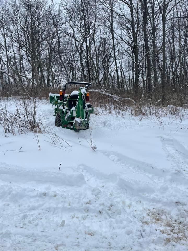 Green tractor with snow blower clearing a snowy path in a wooded area.