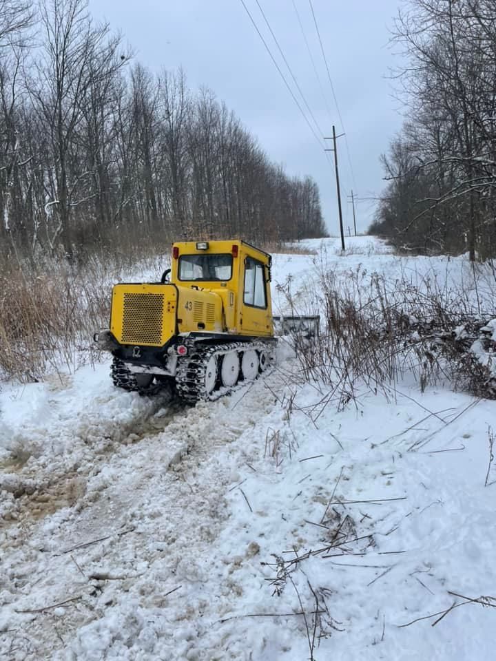 Yellow tracked vehicle plowing snow on a wooded path under power lines.