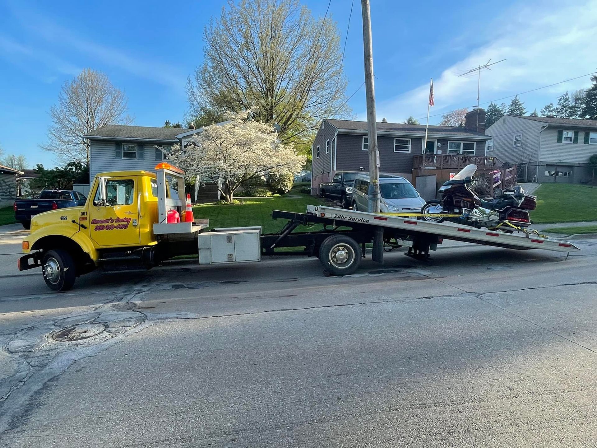 Yellow tow truck on a street, towing a damaged vehicle. Houses and trees in the background.