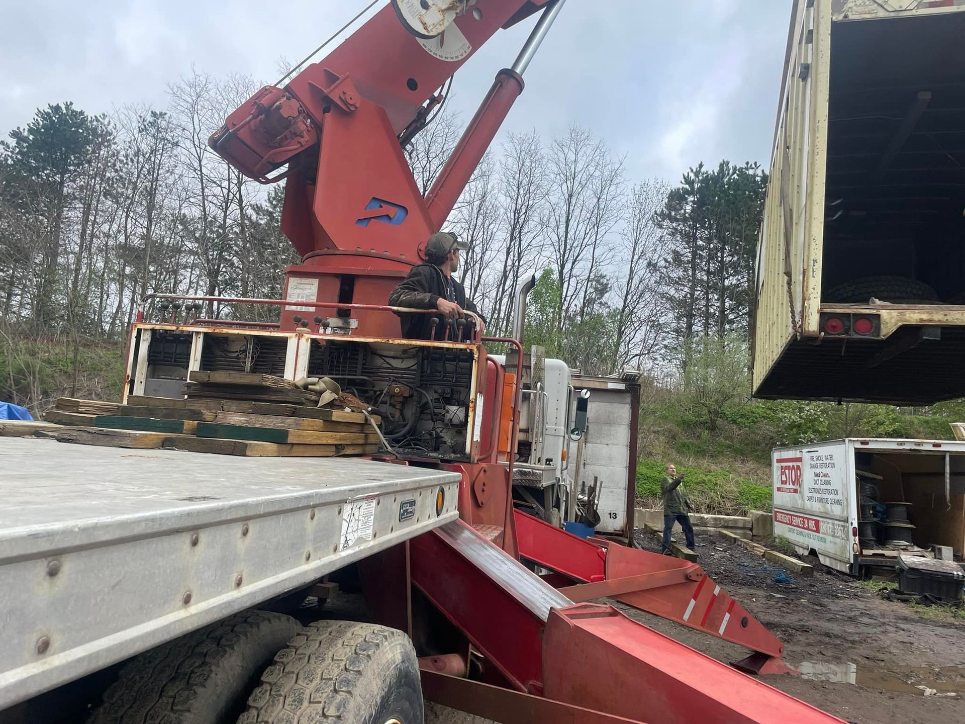 A large crane lifting the side of a trailer. Two people are present, outdoors, in a wooded area.