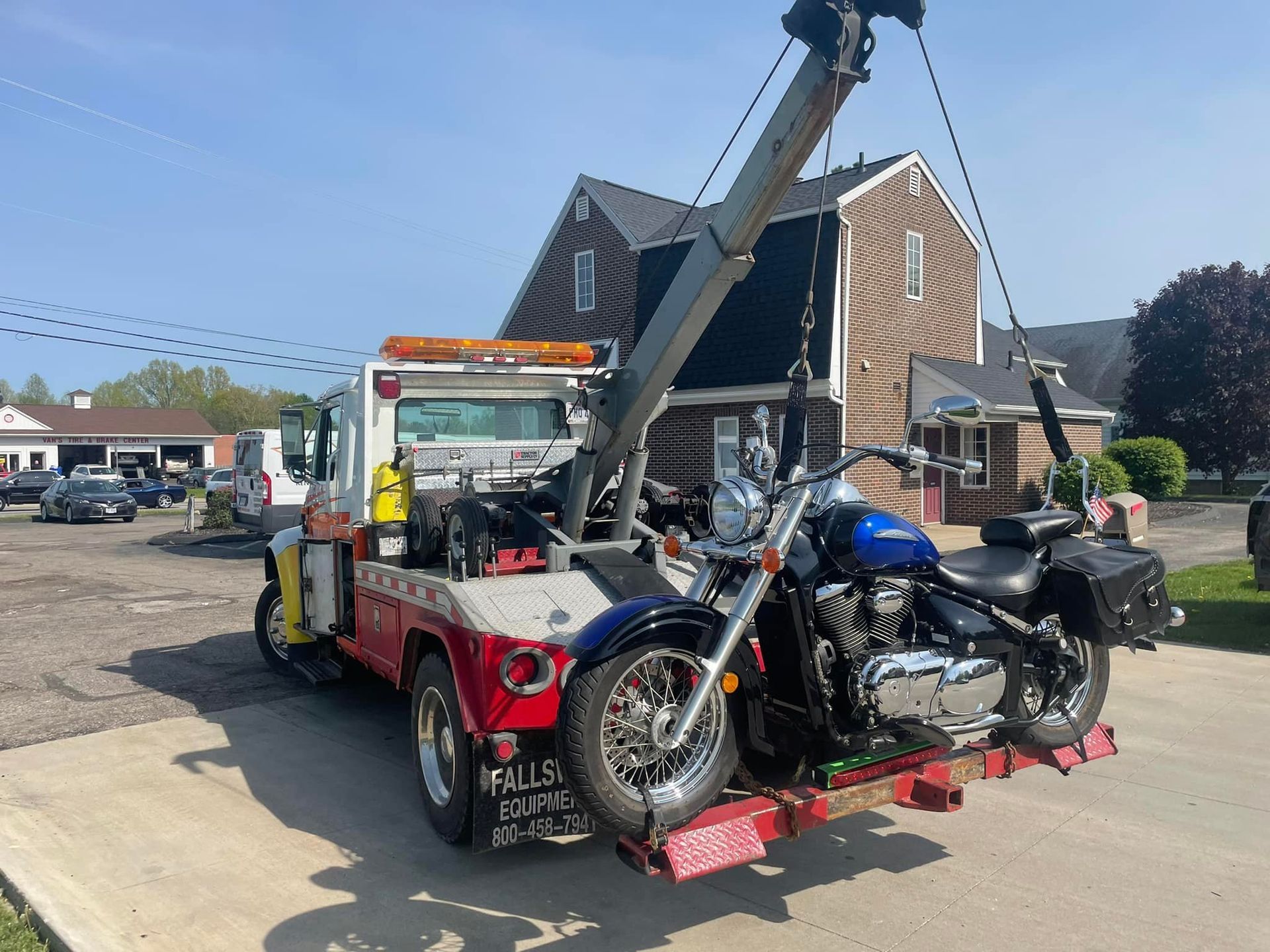 Tow truck with a blue motorcycle being lifted, outside a building on a sunny day.