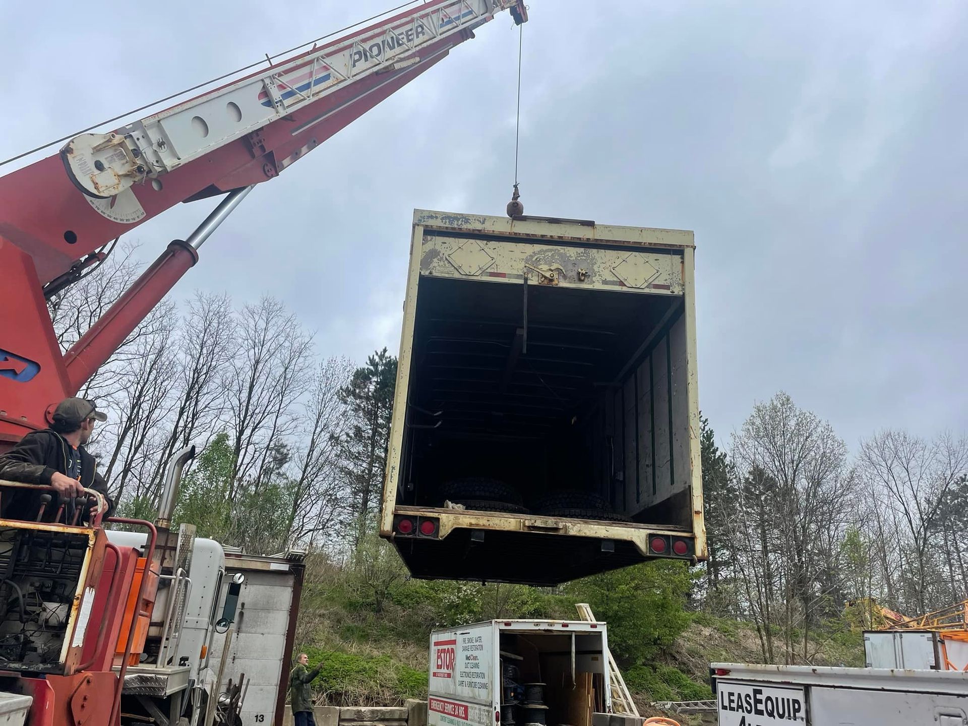 A crane lifting a tan container. A person watches from the left, near a damaged truck.