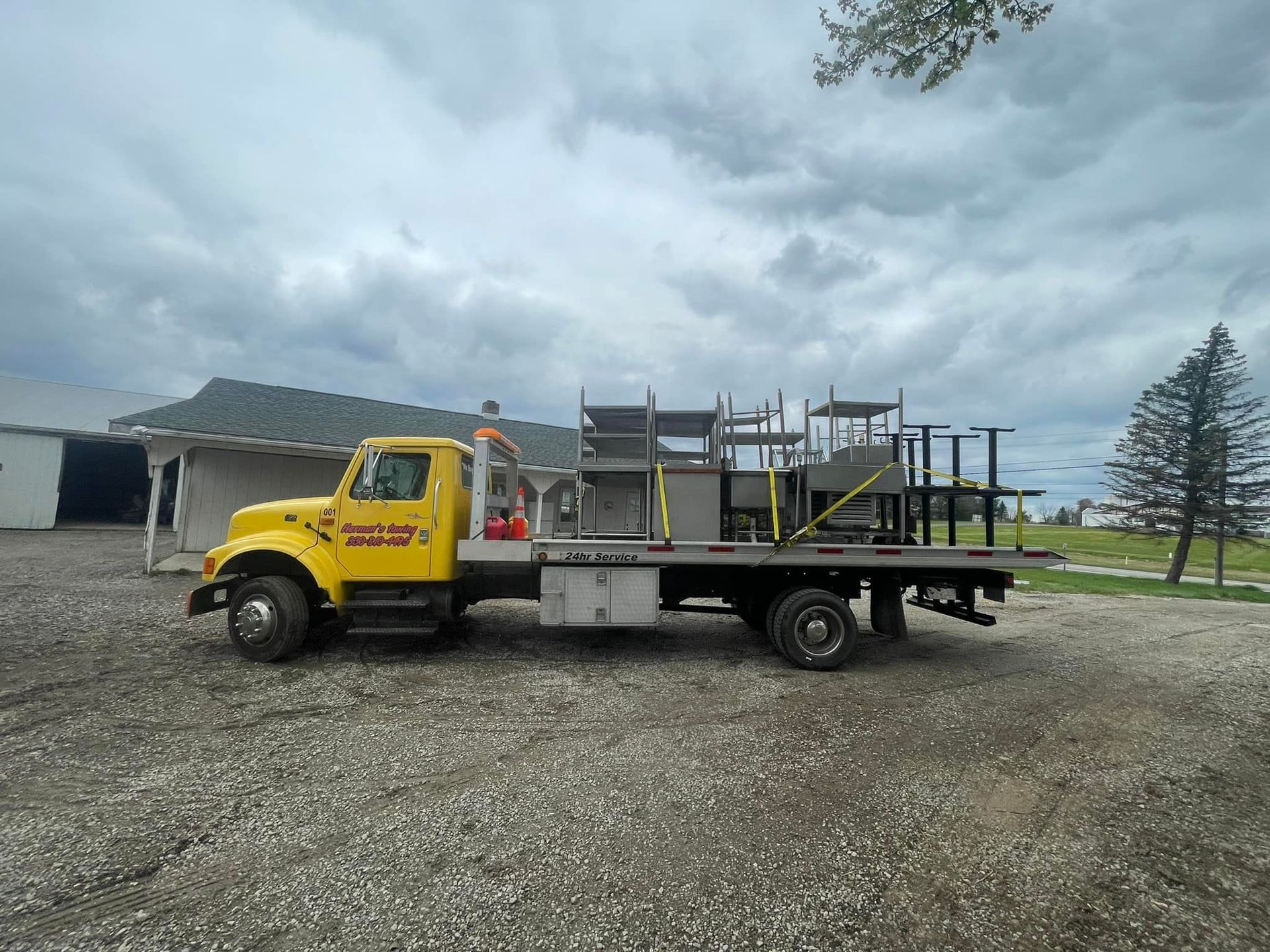 Yellow flatbed truck loaded with metal shelving on gravel driveway under cloudy sky.