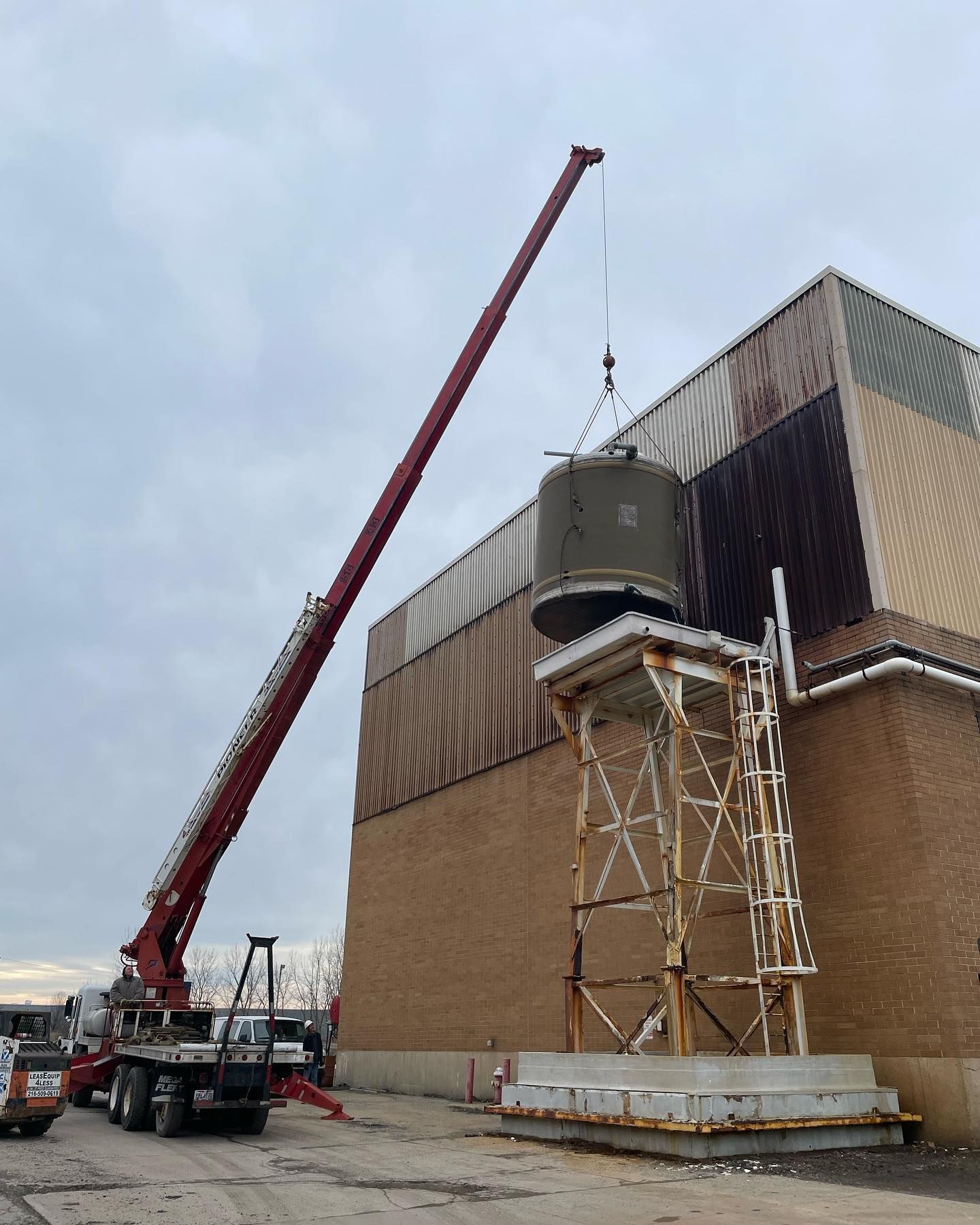 A crane lifts a cylindrical object towards a metal tower on a brick building, under a cloudy sky.