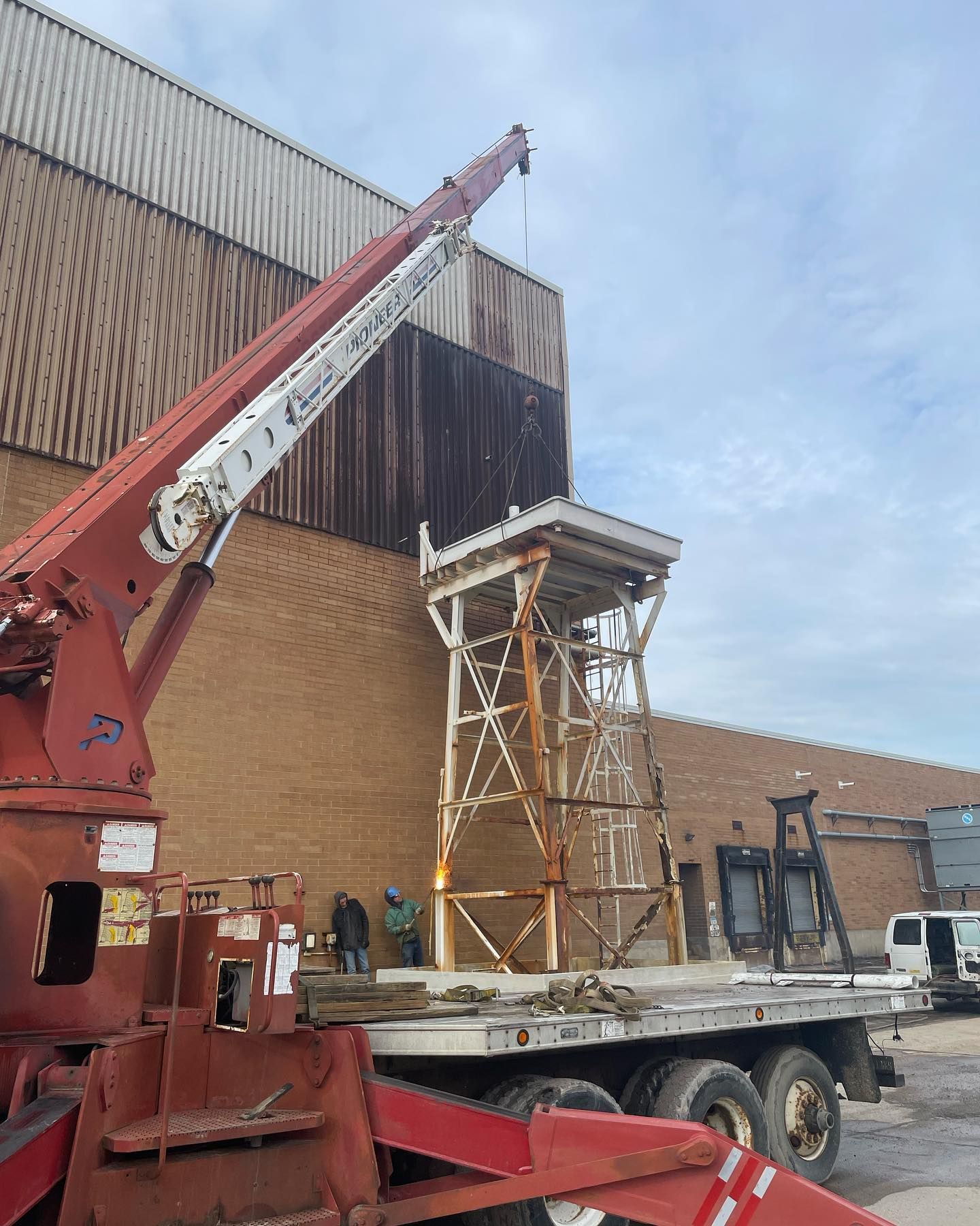 A crane lifting a rusty metal tower onto a flatbed truck near a brick building.