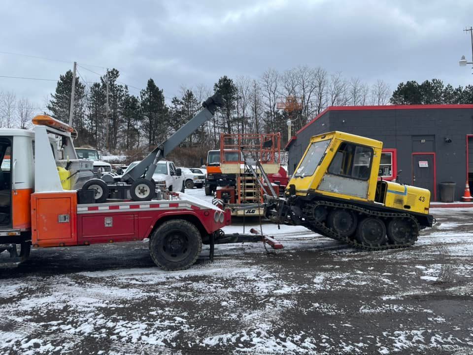 Tow truck loading a yellow snow groomer on tracks; snowy parking lot.