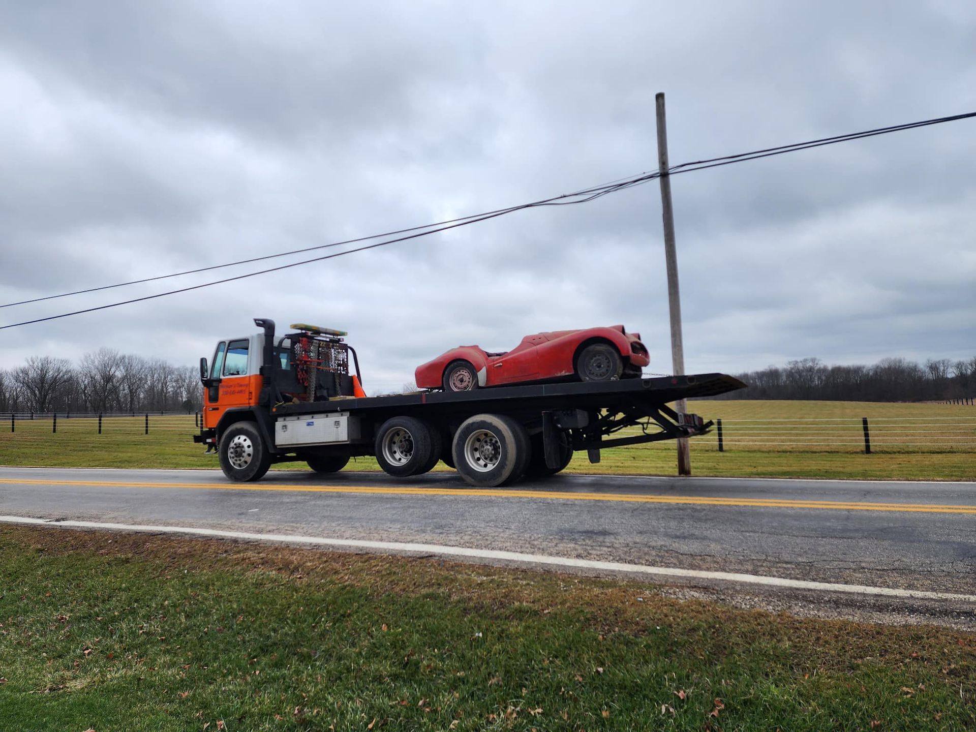 Tow truck carrying a red classic car on a flatbed, driving on a rural road under an overcast sky.