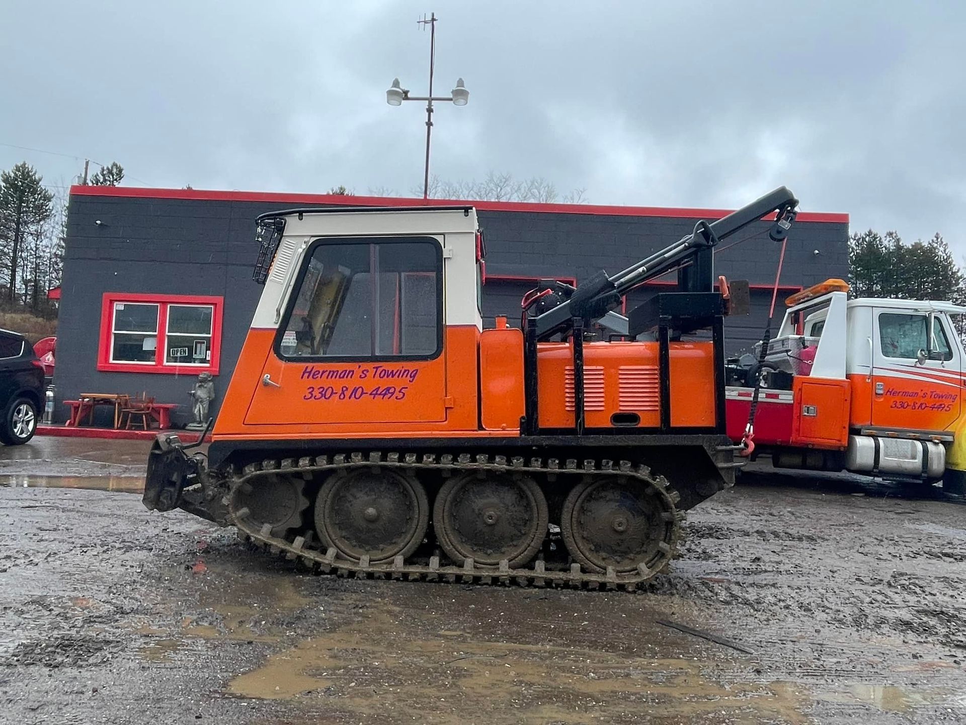 Orange tracked vehicle with a crane next to a building and tow truck.