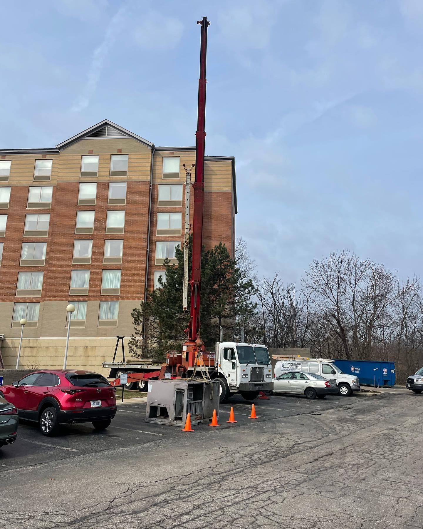 A crane is set up in a parking lot next to a brick building. Cars and a truck are also present.