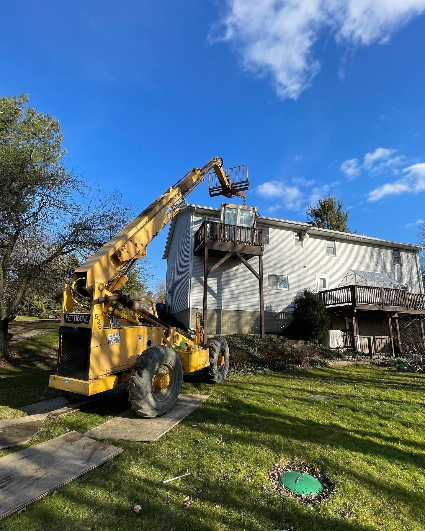 Yellow forklift reaching towards a two-story house with a wooden deck. Blue sky, green grass.