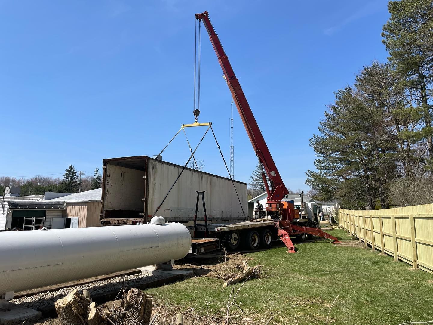 A tow truck lifts a large tree trunk, with a worker atop the truck, in a grassy field under a cloudy sky.