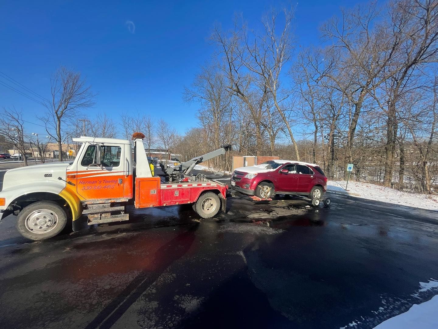 Tow truck towing a red SUV on a snowy day. The tow truck is orange and white, parked on wet asphalt.