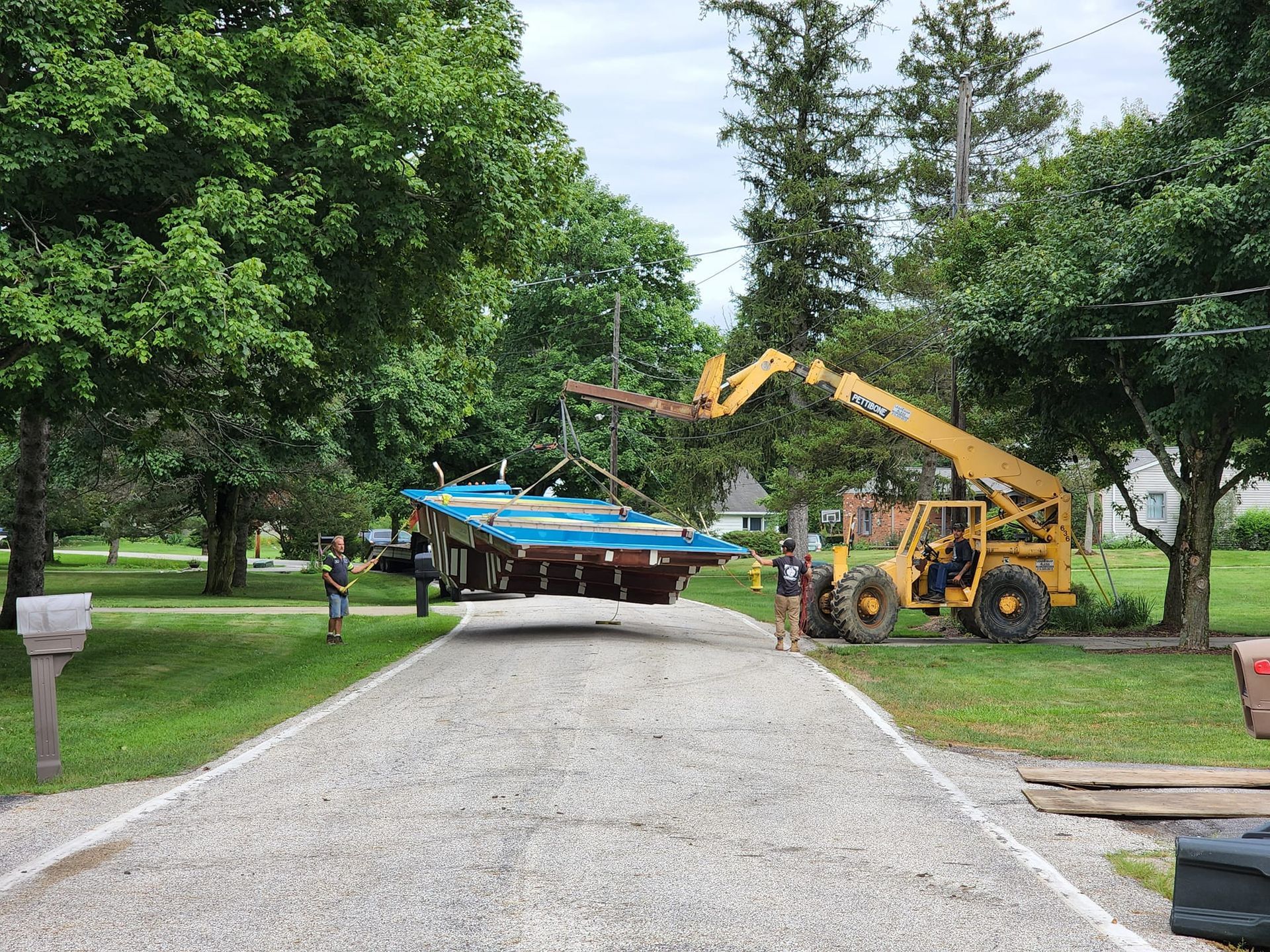 A telehandler lifts a blue platform from a trailer on a gravel driveway, two people watch. Trees surround.