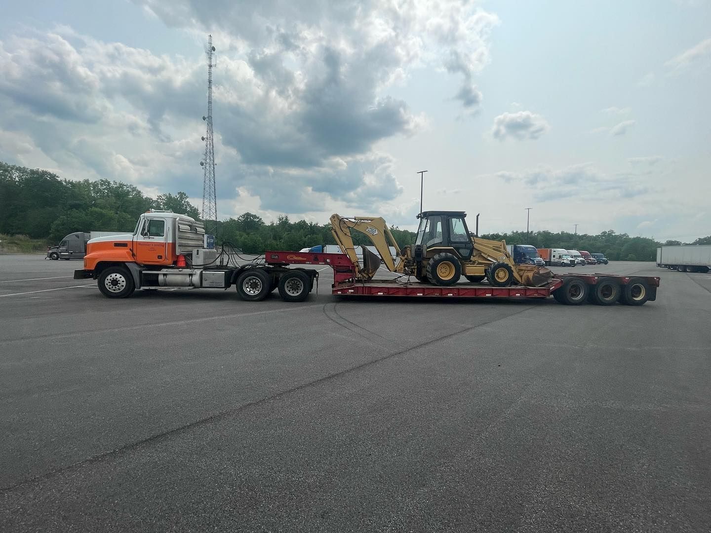 Truck hauling a yellow construction backhoe on a flatbed trailer in a paved lot under a cloudy sky.