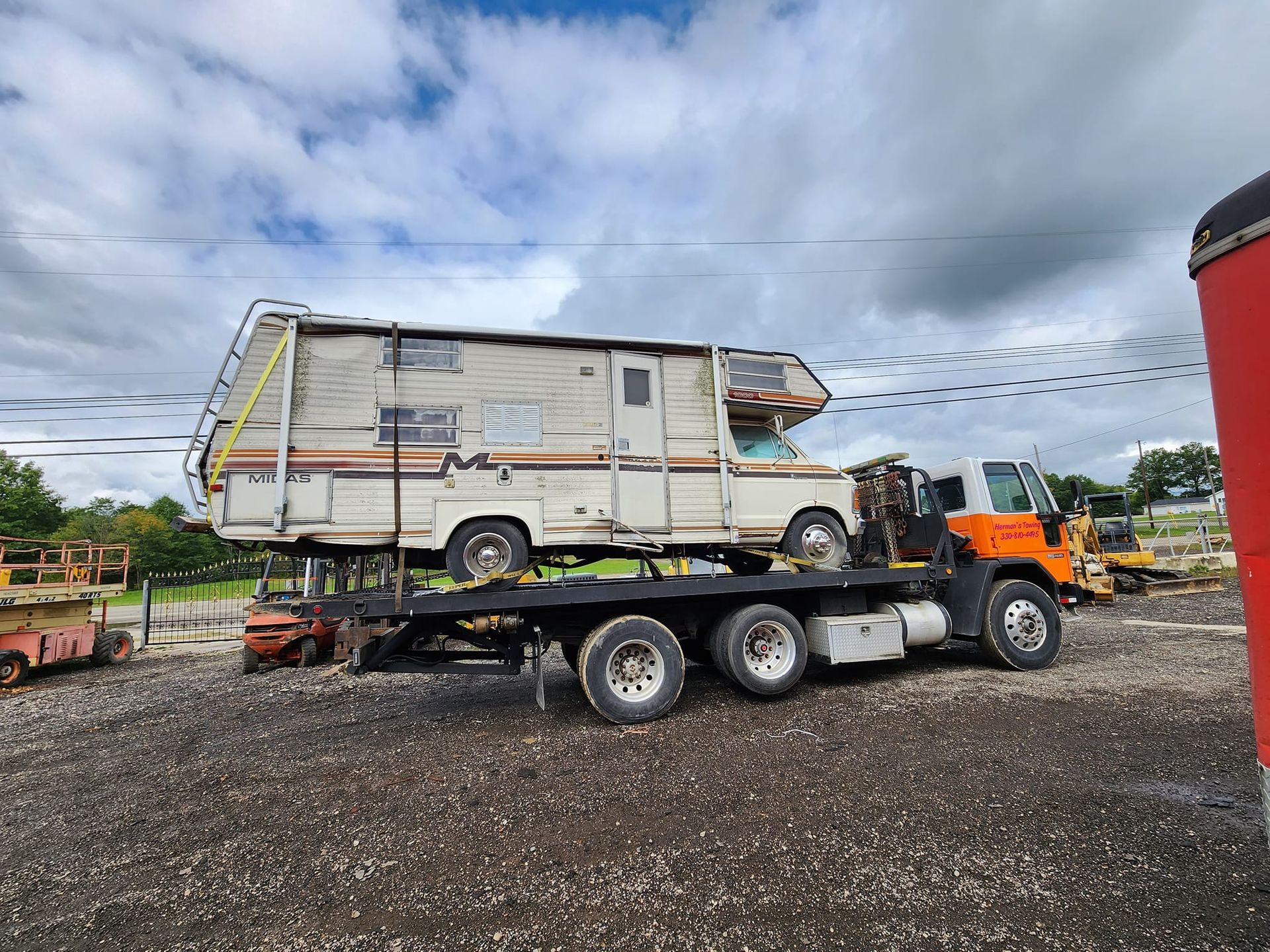 A weathered RV is transported on a flatbed tow truck on a gravel lot under a cloudy sky.