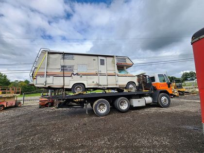 A weathered RV is transported on a flatbed tow truck on a gravel lot under a cloudy sky.