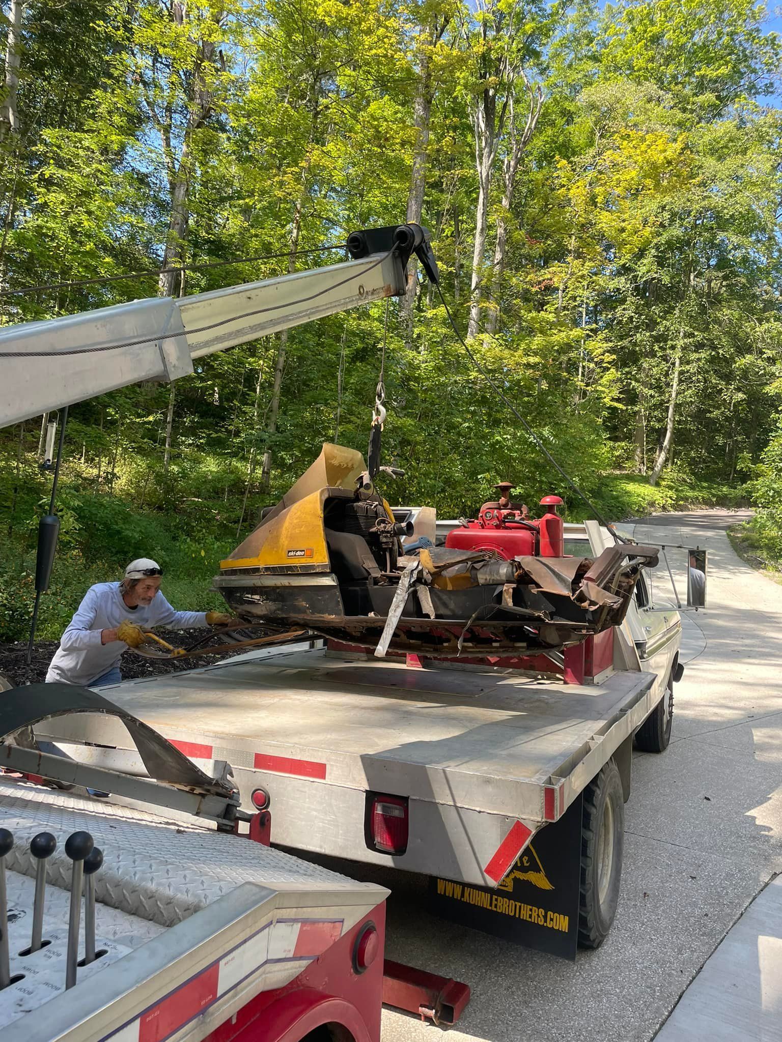 Tow trucks loading a large tree trunk onto a flatbed truck in a field, under a cloudy sky.
